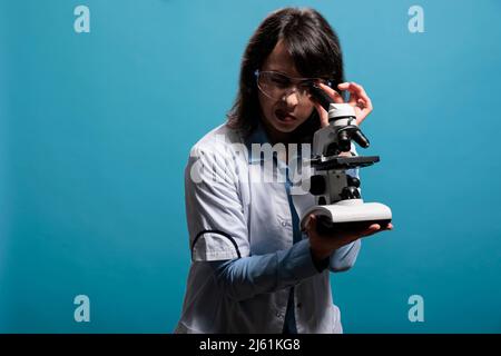 Mikrobiologie-Experte mit modernem Mikroskop, um Viren auf molekularer Ebene auf blauem Hintergrund zu analysieren. Chemiespezialist untersucht chemische Verbindungen in Stoffproben mit dem Mikroskop. Stockfoto
