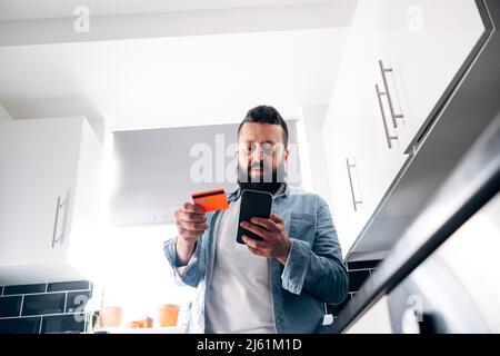 Mann mit Kreditkarte, der zu Hause die Rechnung über das Mobiltelefon bezahlt Stockfoto