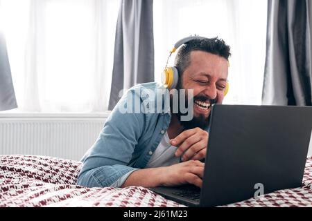Glücklicher Mann mit kabellosen Kopfhörern, der zu Hause mit einem Laptop unterwegs ist Stockfoto