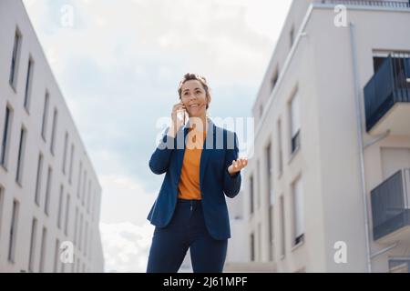 Lächelnde Geschäftsfrau, die am sonnigen Tag vor den Gebäuden mit dem Mobiltelefon spricht Stockfoto