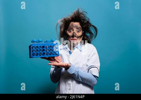 Verrückter Chemiker mit unordentlichen Haaren und schmutzigem Gesicht, der Plastikröhrchen auf dem Rack hält und die Kamera auf blauem Hintergrund anschaut. Lunatic Biochemistry expert mit Stand mit Testflaschen. Studioaufnahme. Stockfoto