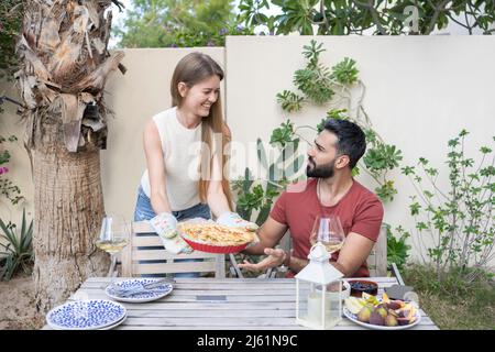 Glückliche Frau, die dem Mann, der am Tisch im Garten sitzt, hausgemachten herzhaften Kuchen zeigt Stockfoto