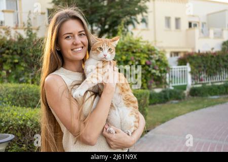 Glückliche Frau mit langen blonden Haaren, die Ingwerkatze im Garten hält Stockfoto