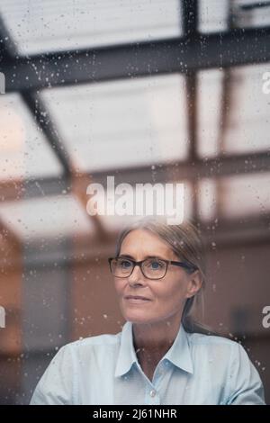 Eine durchdachte Geschäftsfrau, die durch ein Glas Arbeitsplatz gesehen wird Stockfoto