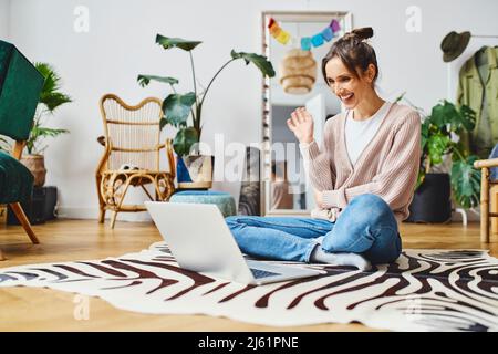 Glücklicher Freiberufler, der mit der Hand durch einen Videoanruf auf einem Laptop mit gekreuzten Beinen zu Hause sitzt Stockfoto