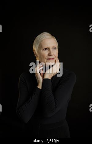 Nachdenkliche ältere Frau mit kurzen blonden Haaren vor schwarzem Hintergrund Stockfoto
