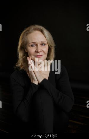Traurige Frau mit blonden Haaren, die vor schwarzem Hintergrund sitzt Stockfoto