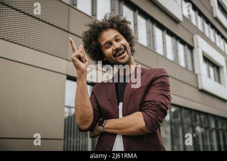 Glücklicher junger Mann mit lockigen Haaren, der vor dem Gebäude zeigt Stockfoto