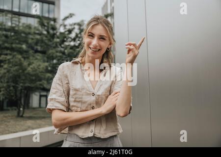 Glückliche junge Frau, die vor einem modernen Gebäude gestikelt wird Stockfoto