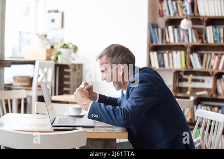 Begeisterter Geschäftsmann, der mit einem Laptop am Tisch sitzt und im Café den Erfolg feiert Stockfoto