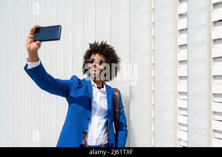Glücklicher Geschäftsmann, der Selfie über das Mobiltelefon macht, das vor der Wand steht Stockfoto