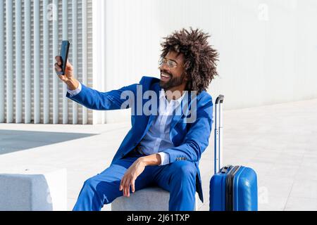 Glücklicher Geschäftsmann mit Koffer, der Selfie über das Mobiltelefon vor der Wand macht Stockfoto
