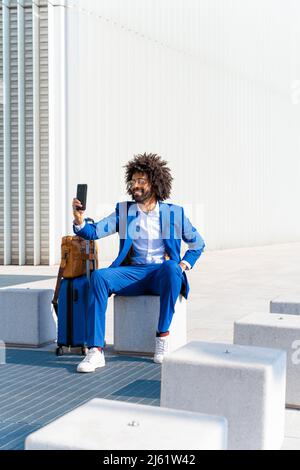 Lächelnder Geschäftsmann mit Gepäck, der Selfie durch ein Smartphone auf einem Betonblock nimmt Stockfoto