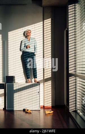 Geschäftsfrau mit Kaffeetasse, die mit der Hand in der Tasche auf dem Sideboard im Büro steht Stockfoto