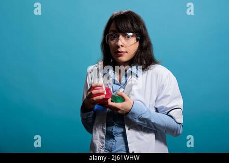 Vorsichtiger, professioneller Chemielabor auf blauem Hintergrund mit Erlenmeyer-Glasgefäßen. Mikrobiologie-Experte, der chemische Becher hält, die mit flüssigen Stoffproben gefüllt sind. Studioaufnahme. Stockfoto