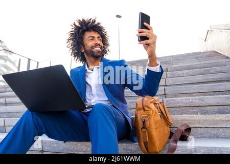 Glücklicher Geschäftsmann mit Laptop, der Selfie über das Mobiltelefon macht und auf der Treppe sitzt Stockfoto