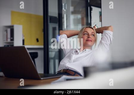 Eine Geschäftsfrau sitzt mit den Händen hinter dem Kopf und einem Laptop am Schreibtisch Stockfoto