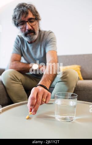 Älterer Mann mit einem Glas Wasser und der Einnahme von Medikamentenpille sitzt zu Hause auf dem Sofa Stockfoto