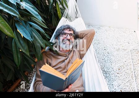 Glücklicher Mann mit Buch, der sich in der Hängematte im Hinterhof entspannen kann Stockfoto