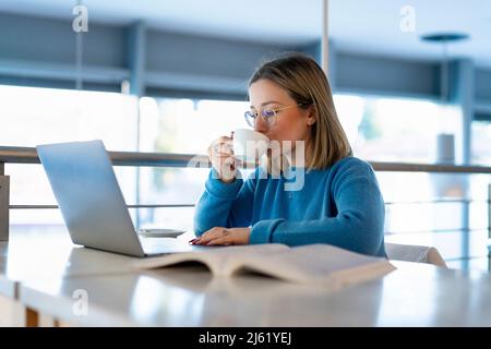 Junge Frau trinkt Kaffee, sitzt mit Laptop und Buch am Tisch Stockfoto