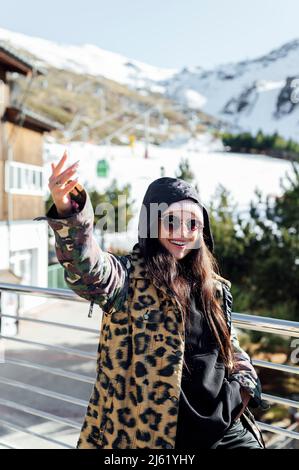 Glückliche Frau in warmer Kleidung, die am sonnigen Tag mit der vom Geländer erhobenen Hand steht Stockfoto