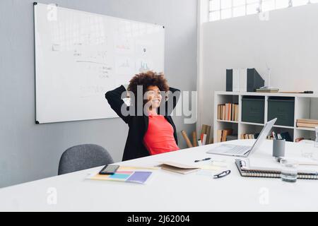 Lächelnde Geschäftsfrau mit Händen hinter dem Kopf, die im Büro am Schreibtisch sitzt Stockfoto