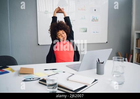 Müde junge Geschäftsfrau, die sich am Schreibtisch im Büro streckt Stockfoto