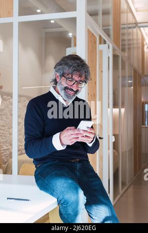 Glücklicher älterer Mann, der zu Hause über das Mobiltelefon im Internet surfen und auf dem Tisch sitzen kann Stockfoto