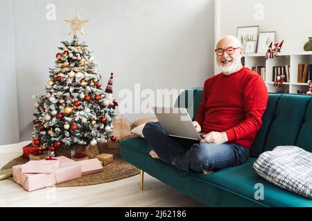 Lächelnder älterer Mann mit Laptop sitzt zu Hause auf dem Sofa Stockfoto