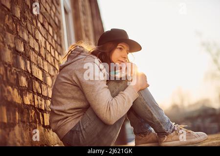 Wartende junge Frau mit Baseballmütze, die auf dem Boden sitzt und die Knie umarmt Stockfoto