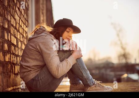 Wartende junge Frau mit Baseballmütze, die auf dem Boden sitzt und die Knie umarmt Stockfoto