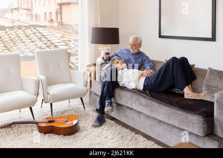 Frau, die auf dem Schoß des Mannes liegt und mit Weinglas auf dem Sofa im Wohnzimmer sitzt Stockfoto