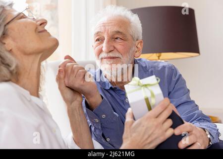 Ein älterer Mann sieht eine Frau, die mit einem Geschenk im Hotelappartement sitzt Stockfoto