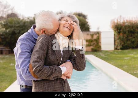 Älterer Mann, der eine fröhliche Frau vor dem Swimmingpool umarmt Stockfoto