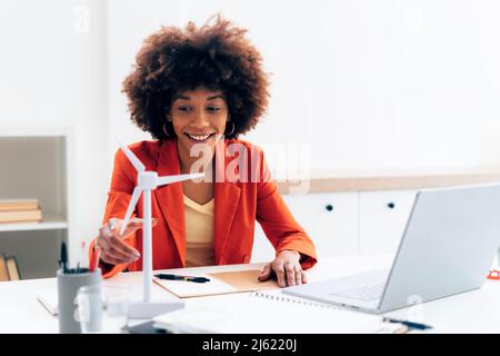 Lächelnde Geschäftsfrau mit Windmühle Modell und Laptop sitzt am Schreibtisch im Büro Stockfoto