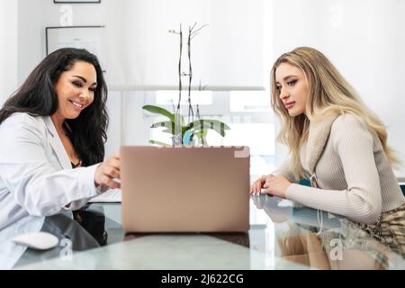 Lächelnder Arzt, der am Laptop mit dem Patienten am Schreibtisch in der medizinischen Klinik diskutiert Stockfoto