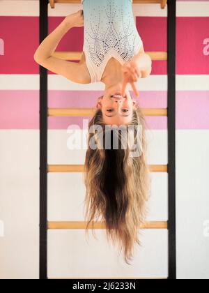 Lächelndes Mädchen, das kopfüber an den Wandleisten hängt und das Friedenszeichen darstellt Stockfoto