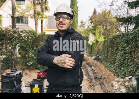 Lächelnder Bauarbeiter, der mit gekreuzten Armen auf der Baustelle steht Stockfoto