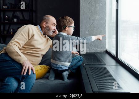 Junge zeigt durch das Fenster von Großvater, der zu Hause auf dem Sofa sitzt Stockfoto