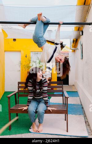 Glückliche junge Frau, die an der Bar über einem Freund hängt, der auf der Bank im Hinterhof sitzt Stockfoto