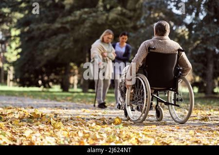 Älterer behinderter Mann, der im Rollstuhl sitzt, vor einer Krankenschwester, die mit einer Frau im Park läuft Stockfoto