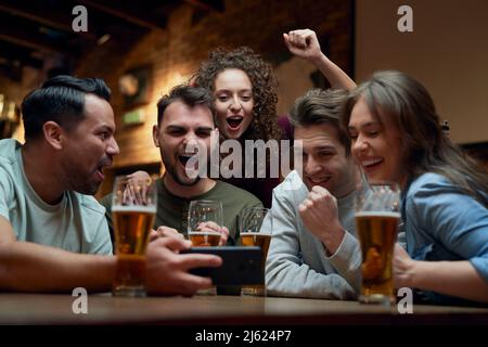 Eine Gruppe fröhlicher Fußballfans, die Bier trinken und ein Spiel auf dem Smartphone in einem Pub sehen Stockfoto