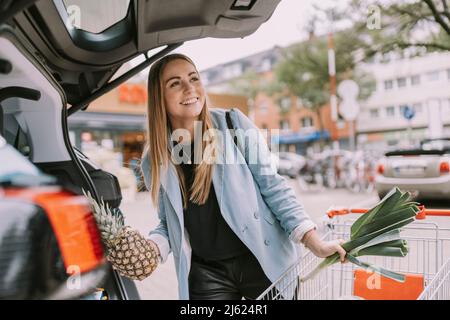 Glückliche junge Frau mit Einkaufswagen, die Lebensmittel in den Kofferraum einlädt Stockfoto