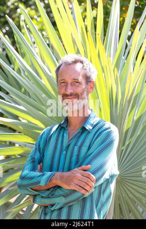 Lächelnder Mann mit gekreuzten Armen, der an sonnigen Tagen vor dem Palmblatt stand Stockfoto
