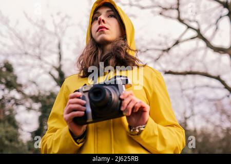 Junge Frau im gelben Regenmantel, die mit der Kamera im Wald steht Stockfoto