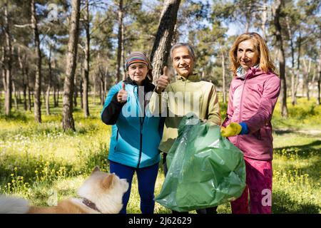 Lächelnder Mann und Frau, die Daumen hoch gestikulieren, von einem Freund, der mit einem Müllbeutel im Wald steht Stockfoto