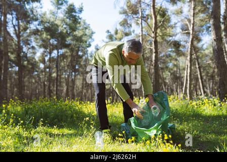 Reifer Mann sammelt Plastikflaschen im Müllbeutel im Wald Stockfoto