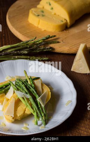 Gerollter Maisbrei mit Spargel und Käseflocken auf weißem Teller und dunklem Holzhintergrund Stockfoto