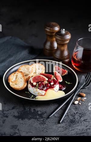 Gebackener Camembert oder Brie-Käse mit Cranberry-Sauce und frischen Feigen auf einem schwarzen Teller, Nahaufnahme Stockfoto