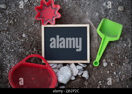 Draufsicht auf eine kleine leere Tafel, die auf einem sandigen Boden liegt und um sie herum Sandboden-Spielzeuge für Kinder in einem konzeptuellen Bild des Sommerurlaubsangebots. Stockfoto